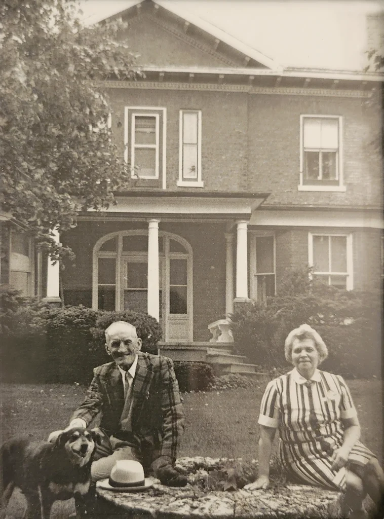 Dog, older man in sport jacket and woman in striped dress seated in front of 2-storey brick house.